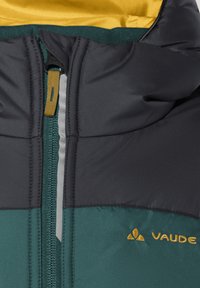 Nahaufnahme des Reißverschlussbereichs einer türkisfarbenen und schwarzen Jacke mit gelbem Reißverschluss-Zipper und gelbem Vaude-Logo, das auf der Brust gestickt ist.