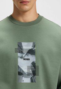 Felpa verde con una stampa grafica di edifici urbani e montagne, con la parola "BOSS" centrata, accentuata da linee nere.