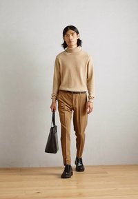 Rjava turtleneck pulover, bež hlače in črne čevlje. Drži črno usnjeno torbo. Gladke teksture, sproščen kroj, minimalističen dizajn.