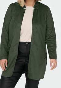 Chaqueta larga de gamuza verde con cuello chal, bolsillos laterales y corte recto. Se lleva sobre una camiseta de color claro y pantalones negros.