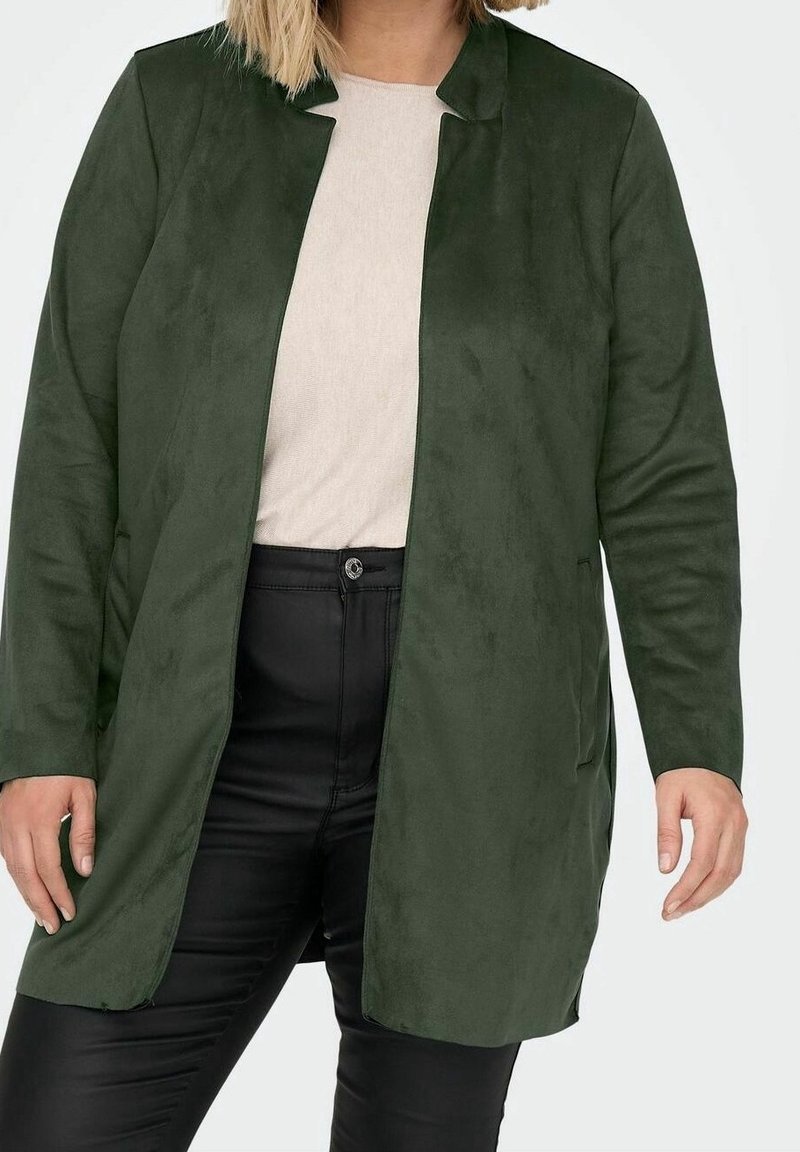 Chaqueta larga de gamuza verde con cuello chal, bolsillos laterales y corte recto. Se lleva sobre una camiseta de color claro y pantalones negros.
