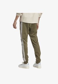 Olivgröna joggerbyxor med tre vita sidostripes, mjukt tyg, åtsittande mudd, kombinerade med vita sneakers och en ljusbeige skjorta.