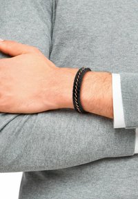 Pulsera de goma negra con un diseño trenzado, que presenta acentos metálicos en color rosa dorado. Lleva puesta en una muñeca sobre un suéter de punto gris.