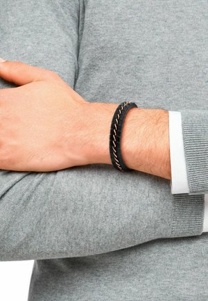 Czarna gumowa bransoletka o plecionym wzorze, z metalowymi różowo-złotymi akcentami. Noszona na nadgarstku, powyżej szarego swetra z dzianiny.