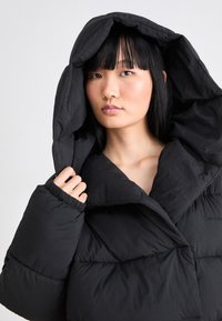 Chaqueta de plumas negra con un amplio cuello, capucha oversized y puños elásticos. Fabricada con un tejido suave y ligero con una textura acolchada.