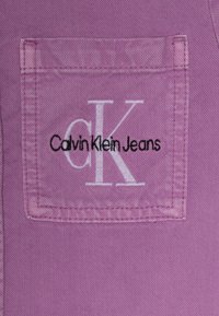 Fioletowa dżinsowa tkanina z prostokątną tylną kieszenią z białym haftem "CK" oraz napisem "Calvin Klein Jeans". Widoczna faktura.