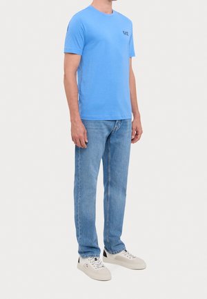 Hellblaues T-Shirt mit kurzen Ärmeln, das einen Rundhalsausschnitt und ein kleines schwarzes Logo auf der linken Brust hat. Kombiniert mit hellblauen Jeans und weißen Sneakers.