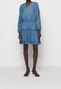 Farkkutunika, jossa on V-pääntie, pitkät hihat ja kuminauha vyötäröllä. Sirot volangit hemman alareunassa. Pehmeä tekstuuri; vaaleansininen väri.