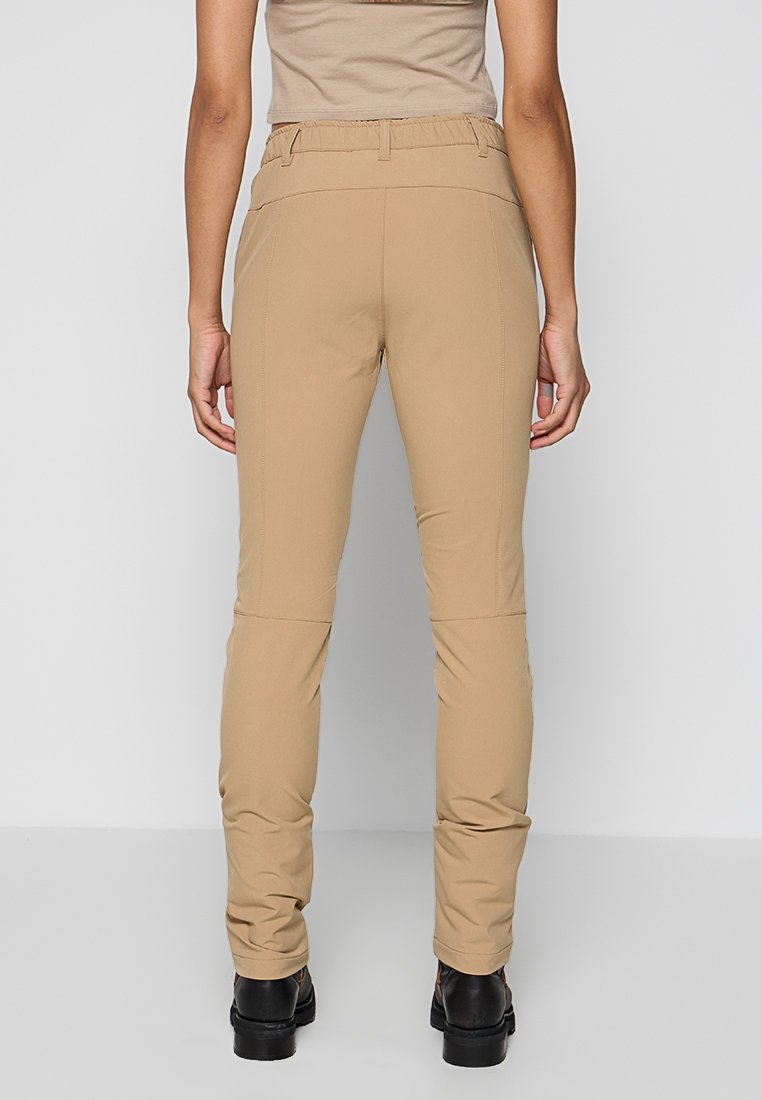 Widok z tyłu osoby w beżowych slim-fit spodniach, beżowej bluzce i czarnych botkach do kostek na jednolitym szarym tle.