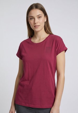 Junge Frau mit glatten braunen Haaren, die ein kurzärmeliges dunkelrosa T-Shirt mit aufgekrempelten Ärmeln und dezentem Text auf der Brust trägt.