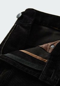 Gros plan sur la ceinture d'un pantalon en velours côtelé noir avec un bouton en métal, une fermeture éclair et une doublure en tissu marron à l'intérieur.