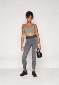Leggings atléticos cinzentos com riscas texturizadas, combinados com um soutien desportivo verde-claro. O modelo segura um kettlebell preto. Ténis pretos completam o look.