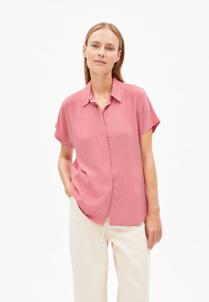 Kurzärmlige, knöpfbare Bluse aus sanftem rosa Stoff. Sie hat einen lässigen Schnitt, einen Kragen und unauffällige Knöpfe. Kombiniert mit cremefarbener Hose.