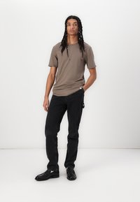 Braunes Kurzarmhemd mit Rundhalsausschnitt, kombiniert mit schwarzen geraden Jeans und schwarzen Schuhen. Lässiges Outfit vor hellem Hintergrund.