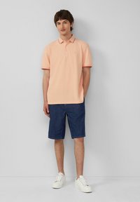 Pfirsichfarbenes Poloshirt mit Kragen und kurzen Ärmeln, kombiniert mit dunkelblauen Denim-Shorts und weißen Turnschuhen. Einfaches Design.