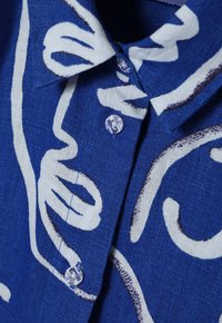 Camicia in lino blu con motivi astratti bianchi, chiusura con bottoni trasparenti e colletto strutturato.