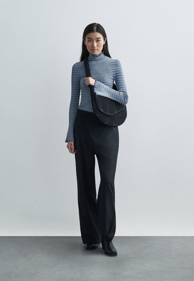 Donna che indossa una maglietta a righe blu con maniche lunghe, pantaloni a gamba larga neri a righe, stivali neri e porta una borsa nera a tracolla, in piedi su un pavimento grigio.