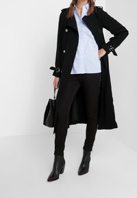 Woman wearing long black coat, light blue button-up shirt, black pants, black heeled boots, holding black handbag, standing in white space.