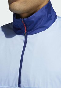 Veste bleu clair avec un col bleu marine, dotée d'une fermeture à demi-zip. Le tissu semble lisse avec une finition durable et un accent rouge sur la fermeture éclair.