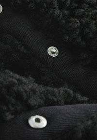 Svart texturerad tyg har en fluffig yta, med silvermetallknappar som kontrasterar mot det släta canvasmaterialet. Anmärkningsvärda lager och sömnadsdetaljer.