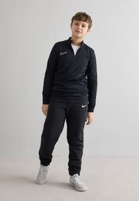 Czarna sportowa koszulka z długim rękawem, z kołnierzem na zamek i logo Nike, w połączeniu z czarnymi spodniami dresowymi z logo Nike oraz szarymi sneakersami.