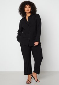 Zwarte op maat gemaakte suit met een knoopjasje met een kraag en twee voorzakken, gecombineerd met rechte broekspijpen en zwarte hakken.