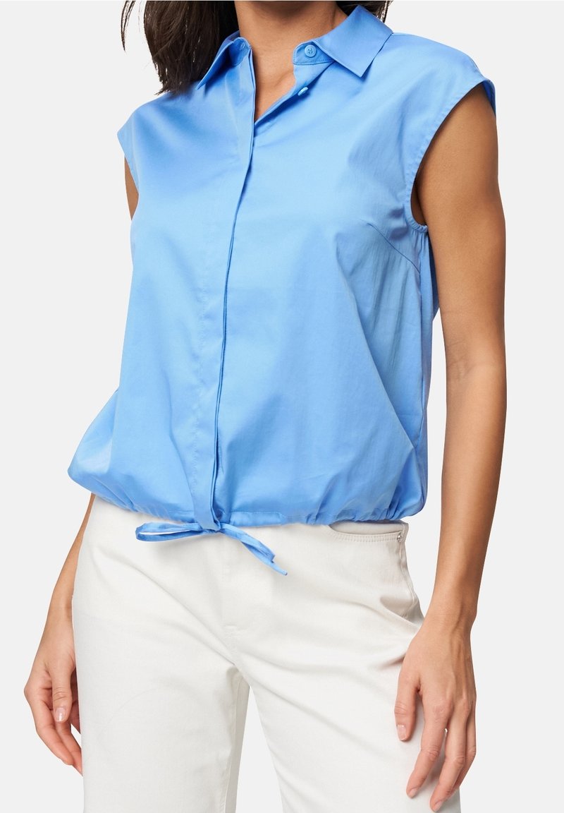 Blouse sans manches bleu clair avec col, boutonnage à l'avant et taille froncée avec lien d'ajustement. Tissu lisse et brillant.