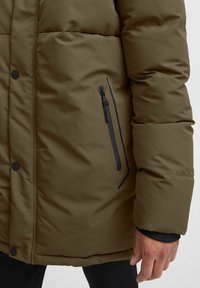 Giacca puffer verde oliva con texture trapuntata, dotata di una tasca con zip nera e bottoni a pressione lungo la parte anteriore.