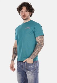 T-shirt in cotone teal con collo rotondo, caratterizzato da testo ricamato in rosa sul lato sinistro del petto. Abbinato a jeans in denim strappati con dettagli in catena.