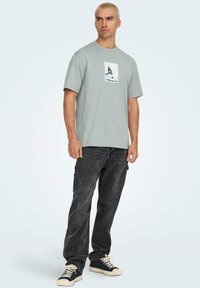 T-shirt di cotone verde chiaro con stampa grafica al centro, abbinata a jeans denim scuri e scarpe da ginnastica nere con suole crema.