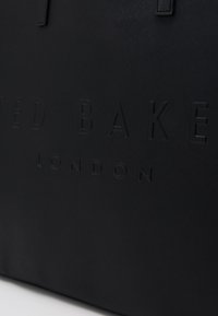 Czarna torba tote z sztucznej skóry z tłoczonym napisem "Ted Baker London". Posiada solidne uchwyty, teksturowaną powierzchnię oraz zorganizowany prostokątny kształt.