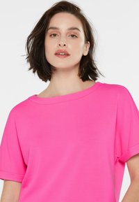 Frau mit kurzem, dunklem Haar, die ein hellrosa, weit geschnittenes Oberteil trägt und selbstbewusst in die Kamera blickt, vor einem schlichten weißen Hintergrund.