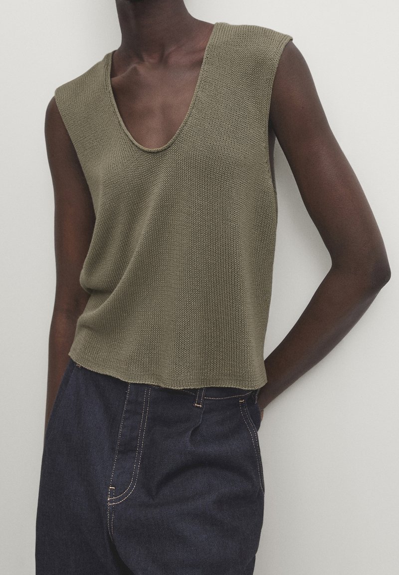 Olive green sleeveless knit top with a deep V-neck, featuring a relaxed fit and raw edge hem. Paired with dark blue denim trousers.
