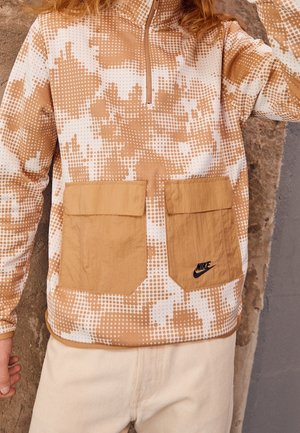 Osoba ubrana w beżowo-biały pikselowy sweter Nike z dwoma przednimi kieszeniami i kołnierzem na pół zamka, stojąca na tle fakturowanej ściany.