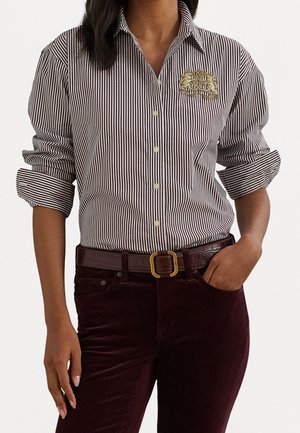 Femme portant une chemise à rayures verticales avec un écusson brodé en or sur la poitrine, assortie d'un pantalon foncé et d'une ceinture marron.