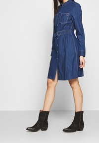 Denim-paita mekko pitkillä hihoilla, napit edessä, kaksi rintataskua ja säädettävä vyö. Tumma sininen väri, jossa on hienovaraisia ompelutarkkuuksia.