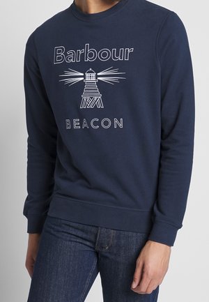 Homme portant un sweat-shirt bleu marine avec un motif phare blanc et le texte « Barbour Beacon », associé à un jean bleu foncé.