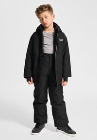 Svart vadderad jacka med dragkedja, fast huva och logodetalj, parat med svarta snöbyxor. Modell iklädd grå tröja och svarta sneakers.