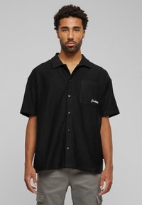 Black short-sleeve shirt made of textured fabric, featuring a collared neckline, front pocket, and logo embroidery on the pocket.