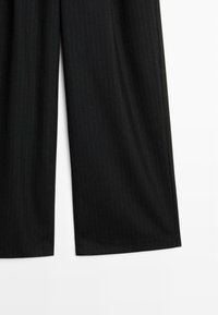 Gros plan sur un pantalon noir à jambes larges, avec de subtiles rayures verticales, montrant les bas des jambes et l'ourlet sur fond blanc.