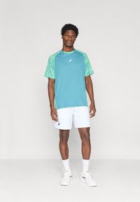 Τυρκουάζ αθλητική μπλούζα με πράσινους διακοσμητικούς μανίκους, συνδυασμένη με λευκά σορτς. Περιλαμβάνει λογότυπο στο στήθος, κανονική εφαρμογή και ελαφρύ ύφασμα.