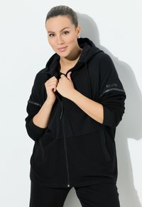 Sudadera con cremallera negra de textura suave, con capucha, mangas acanaladas, detalles plateados y un logo impreso en la manga.