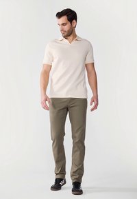 Kremna polo majica s kratkimi rokavi in rebrasto teksturo, kombinirana z olivno zelenimi ozkimi hlačami in črnimi supergami z belimi poudarki.