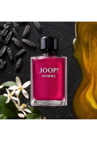 Rosa glasparfymflaska med svart lock, märkt "JOOP! HOMME." Omgiven av svarta frön, vita blommor och en gyllene vätska på en mörk yta.