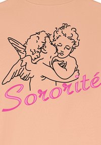 Persiku krāsas T-krekls ar melnu līniju zīmējumu, kurā attēloti divi eņģelīši, kas skūpsta, ar vārdu "Sororité" rakstītu treknajiem rozā rakstiem zemāk.