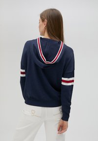 Marinblå huvtröja med röda och vita randiga detaljer, mjuk textur, avslappnad passform och huva. Passar bra med vita jeans med hjärtapplikationer.