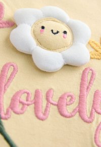 Fleur en tissu doux avec un visage souriant et des joues roses au-dessus du mot brodé en rose "lovely" sur un fond textile jaune clair.