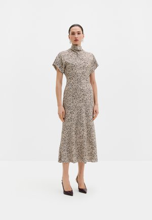 Vrouw draagt een beige midi-jurk met zwart gespikkeld patroon, korte mouwen, hoge halslijn en donkere puntige hoge hakken, staand op een witte achtergrond.