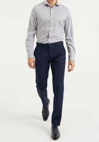 Man som går framåt iförd en ljusgrå knapp-up skjorta, marinblå kostymbyxor och svarta dressade skor mot en vit bakgrund.