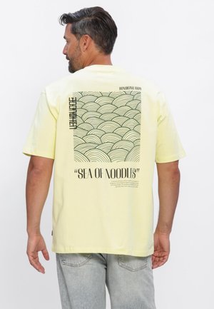 Mand med mørkt hår og skæg iført lysegul t-shirt med sort bølgemønster og teksten "SEA OF NOODLES" på ryggen, kombineret med lysegrå jeans.